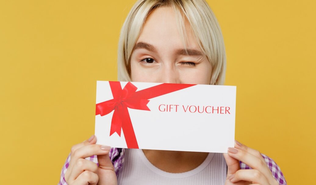 Close up young fun blonde woman 20s wear pink tied shirt white t-shirt hold cover mouth with gift certificate coupon voucher card for store wink isolated on plain yellow background studio portrait.