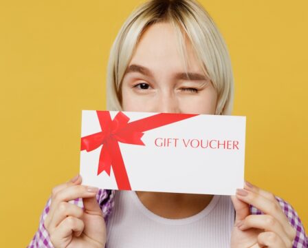 Close up young fun blonde woman 20s wear pink tied shirt white t-shirt hold cover mouth with gift certificate coupon voucher card for store wink isolated on plain yellow background studio portrait.